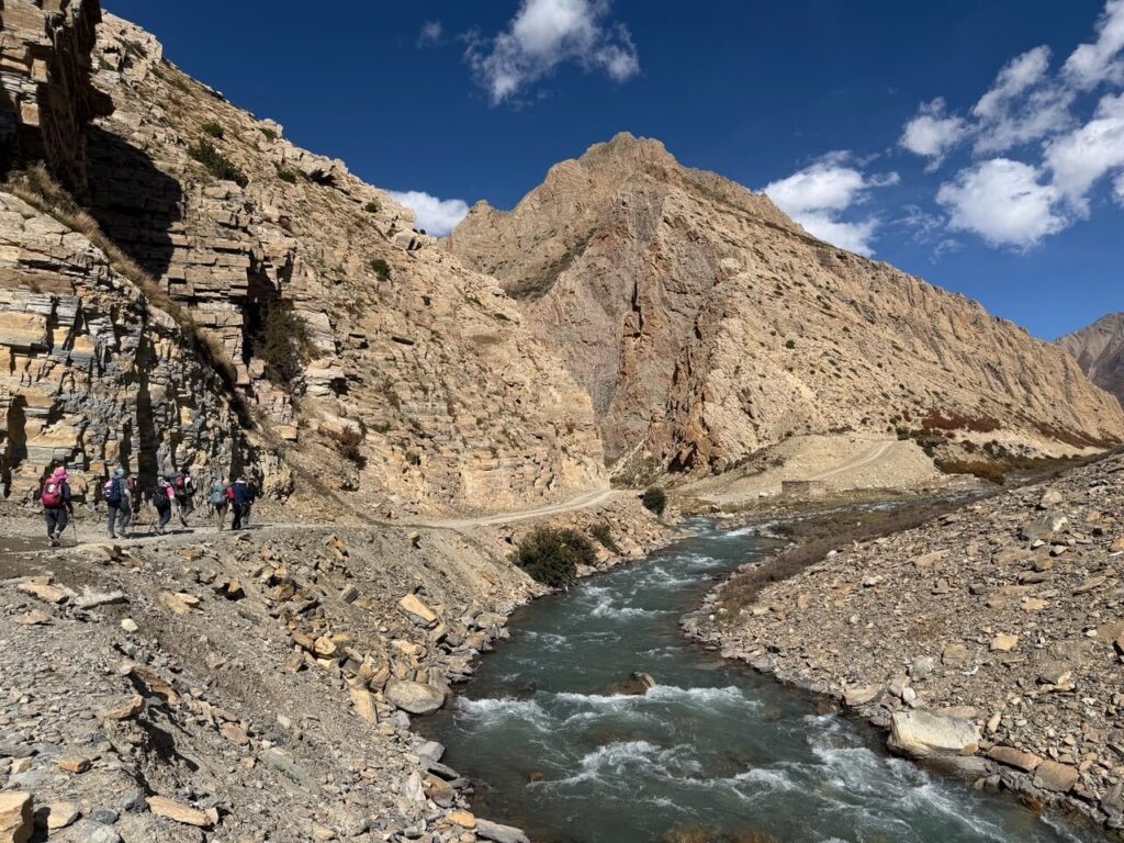 Trekking group walking along a turquoise river below rugged cliffs on the Lower Dolpo trek in Nepal